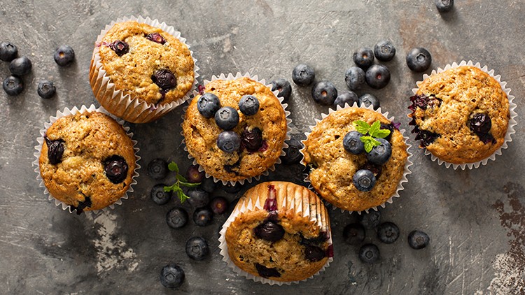 Sechs Muffins mit Blaubeeren und Papierförmchen liegen auf einer grauen Oberfläche, dazwischen frische Blaubeeren.
