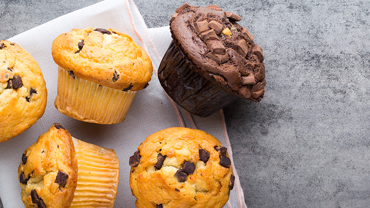 Fünf Muffins, vier mit Schokostückchen und ein dunkler Schokomuffin, liegen auf einem Tuch, Hintergrund in Betonoptik.