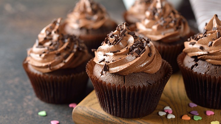 Mehrere Schokoladen-Muffins mit Schokoladencreme und Schokoladenstreuseln, dekoriert mit bunten Herzstreuseln auf Holz.
