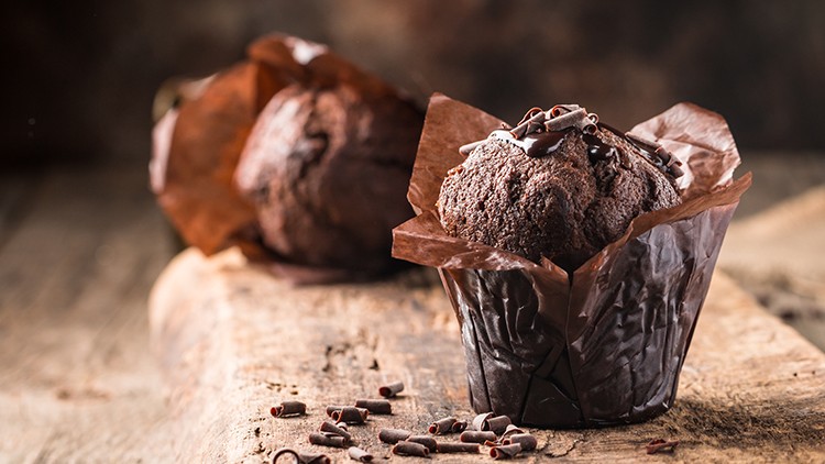 Braune Schokoladenmuffins mit Schokoraspeln auf rustikalem Holzuntergrund, einer im Vordergrund und einer unscharf im Hintergrund.