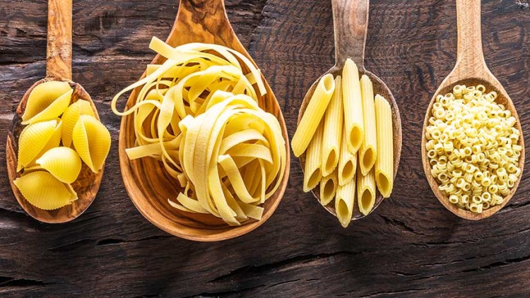 Sieben Holzlöffel mit verschiedenen ungekochten Nudelsorten wie Hörnchen, Farfalle, Spaghetti und Penne auf Holzuntergrund.