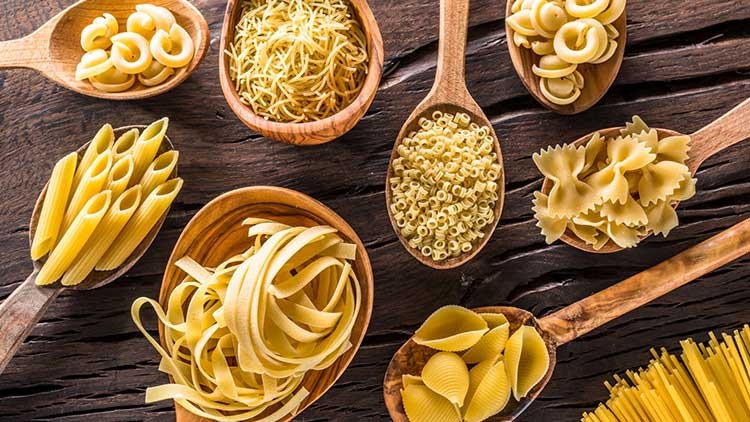 Holzlöffel mit unterschiedlichen ungekochten Nudelsorten wie Farfalle, Penne, Spaghetti und Muscheln auf dunklem Holzuntergrund.