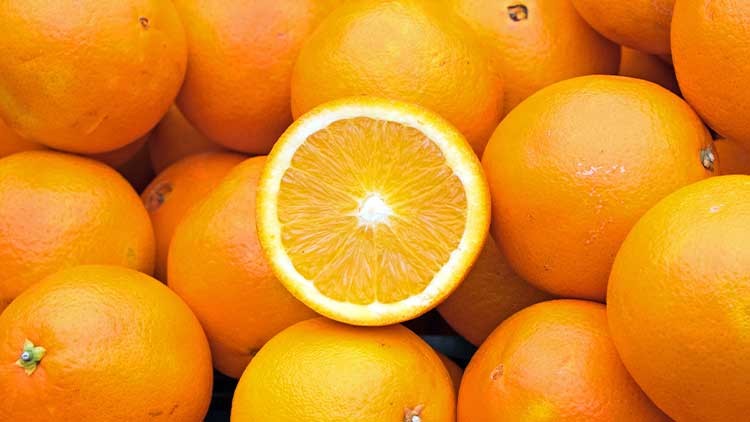 Viele ganze Orangen liegen dicht beieinander, eine davon ist aufgeschnitten und zeigt das saftige Fruchtfleisch.
