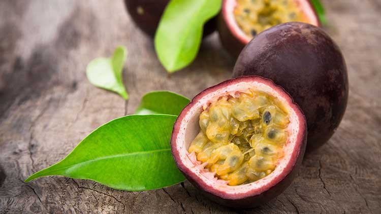Eine aufgeschnittene Passionsfrucht mit gelbem Fruchtfleisch und schwarzen Kernen liegt neben einer ganzen Frucht und grünen Blättern.