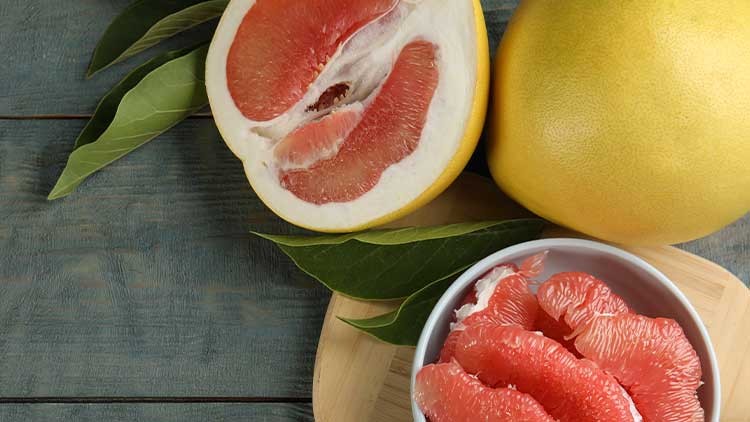 Eine halbierte und eine ganze Grapefruit mit rosa Fruchtfleisch, daneben geschälte Grapefruit-Stücke in einer weißen Schale.