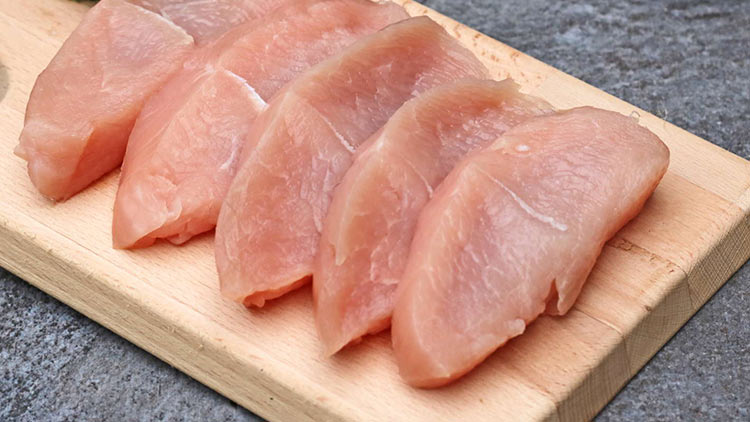 Fünf rohe Hähnchenbrustfilets liegen nebeneinander auf einem hellen Holzbrett, dekoriert mit grüner Petersilie.