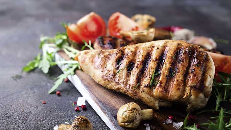 Ein gegrilltes Hähnchenfilet liegt auf einem Holzbrett mit Tomaten, Champignons, Rucola, Pfefferkörnern und Kräutern.