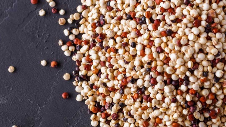 Haufen aus roten, weißen und schwarzen Quinoa-Körnern liegt auf dunklem Steinuntergrund, einige Körner sind verstreut.