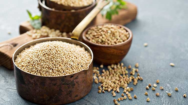 Helle, rohe Quinoa-Körner in einer Kupferschale, daneben zwei Holzschalen mit Quinoa und Dekoration auf grauem Untergrund.
