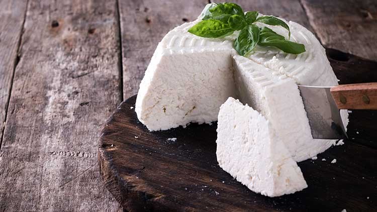Ein runder, weißer Frischkäse mit einem Stück davon abgeschnitten liegt auf einem dunklen Holzbrett, garniert mit Basilikum.