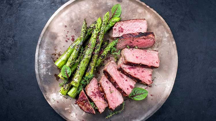 Auf einem Teller liegen gegrillte grüne Spargelstangen und mehrere Scheiben rosa gebratenes Fleisch, dekoriert mit Kräutern.