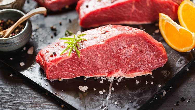 Ein rohes Rindersteak mit Rosmarinzweig liegt auf schwarzer Fläche, daneben Meersalz, Pfeffer und Orangenscheiben.