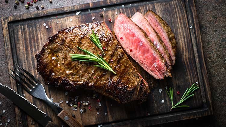 Ein gegrilltes Steak und drei aufgeschnittene Stücke liegen auf einem Holzbrett, dekoriert mit Rosmarin und grobem Salz.