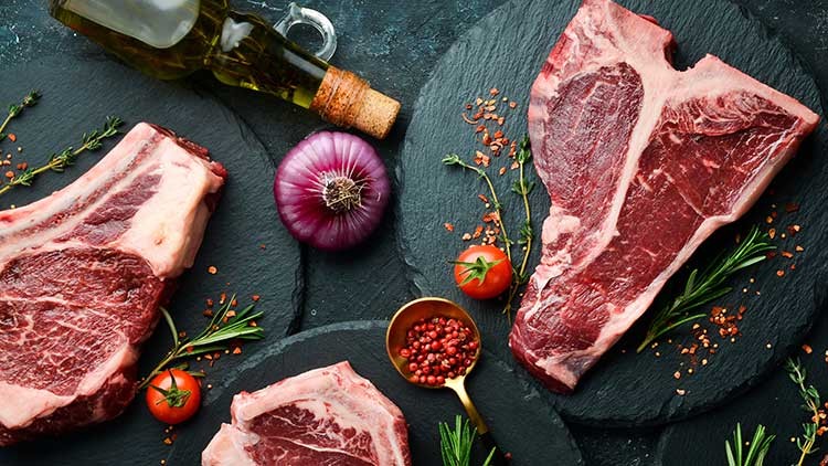 Drei rohe Steakstücke, rote Zwiebel, Kirschtomaten, Rosmarinzweige, Gewürze und eine Flasche Öl auf dunklem Untergrund.