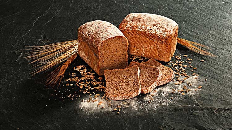 Zwei dunkle Brotlaibe, einer angeschnitten, mit drei Scheiben, Weizenähren und Körnern auf dunklem Untergrund.