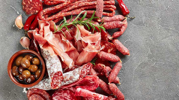 Verschiedene Wurstsorten mit Serranoschinken, Oliven, Knoblauch und Chili auf dunklem Untergrund, rechts eine freie graue Fläche.