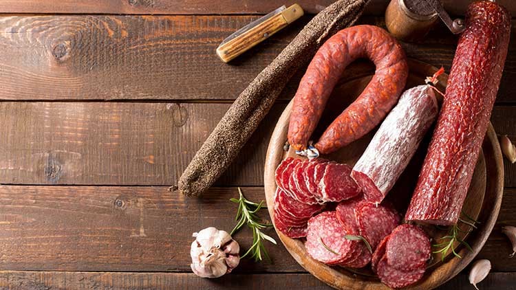 Verschiedene Salamisorten, geschnitten und am Stück, liegen auf einem Holzbrett mit Kräutern, Knoblauch und einem Taschenmesser.