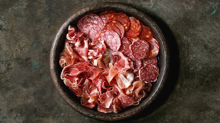 Runde Schale mit verschiedenen Scheiben von Salami, Schinken und anderen Wurstsorten auf dunklem Untergrund.