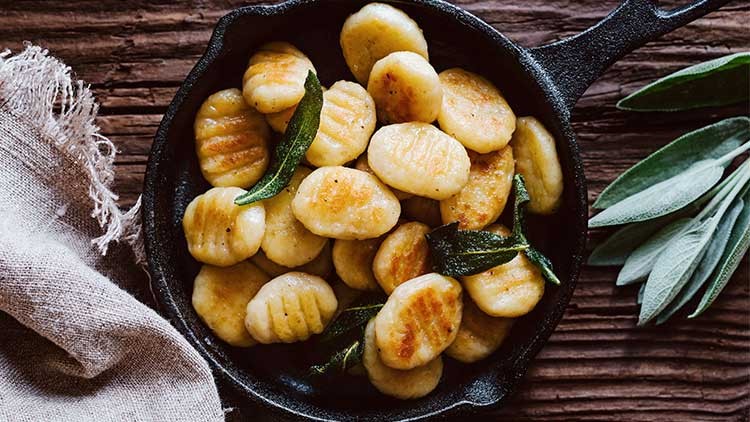 Goldbraun gebratene Gnocchi mit frischen Salbeiblättern in einer schwarzen Pfanne auf rustikalem Holzuntergrund.