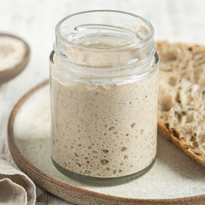 Ein offenes Glas mit Sauerteig, daneben Scheiben helles Brot auf einem Teller, ein Löffel und ein Tuch liegen daneben.