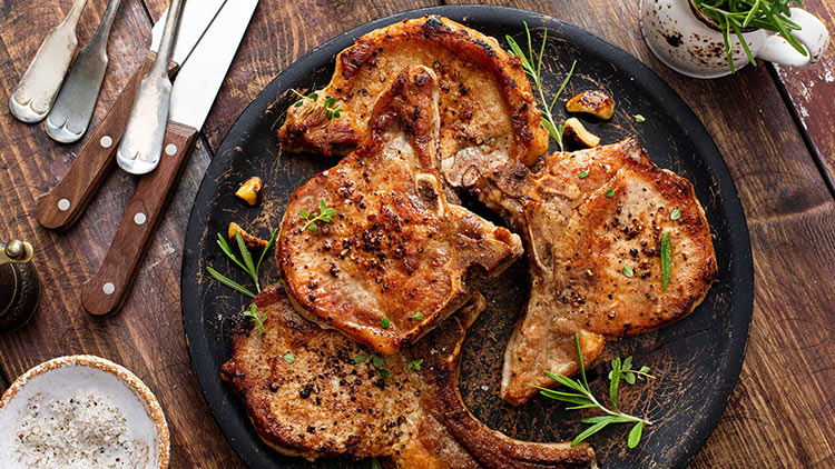 Fünf gebratene Schweinekoteletts mit Kräutern und Gewürzen auf einer schwarzen Pfanne, daneben Holzbesteck auf Holztisch.