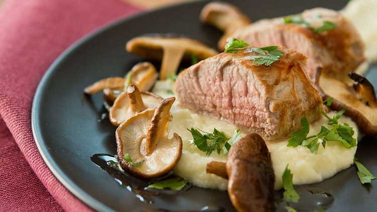 Zwei Scheiben gebratenes Fleisch auf Kartoffelpüree mit Pilzen und Petersilie auf einem schwarzen Teller.