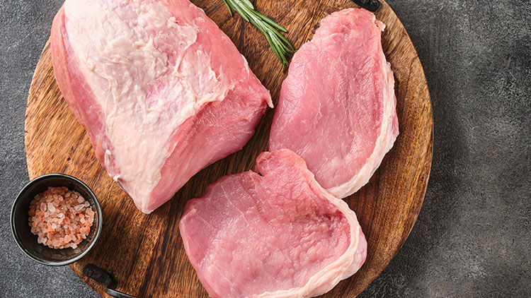 Ein Holzbrett mit drei Stück rohem Schweinefleisch, einem Bündel Rosmarin und einer Schale mit rosa Salz auf dunklem Untergrund.