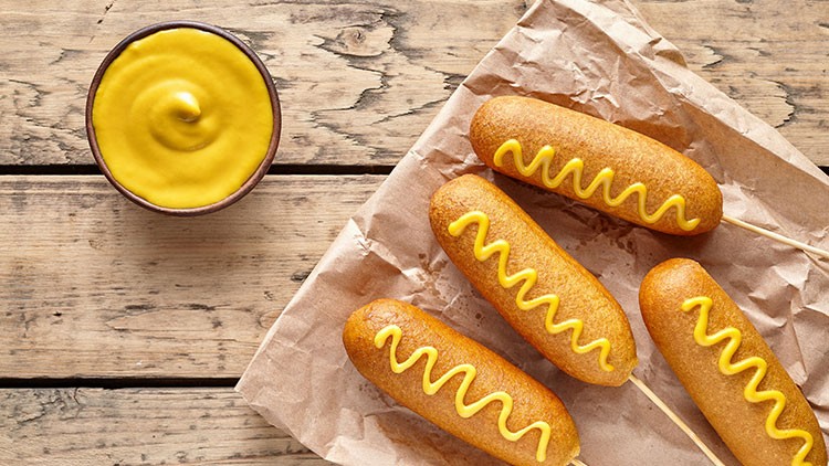 Vier gebackene Corn Dogs mit gelbem Senf darauf, liegen auf braunem Papier neben einer Schale mit Senf auf Holztisch.