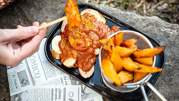 Tablett mit Currywurst auf Baguettescheiben und einem Sieb mit Kartoffelecken, darunter bedrucktes Papier.