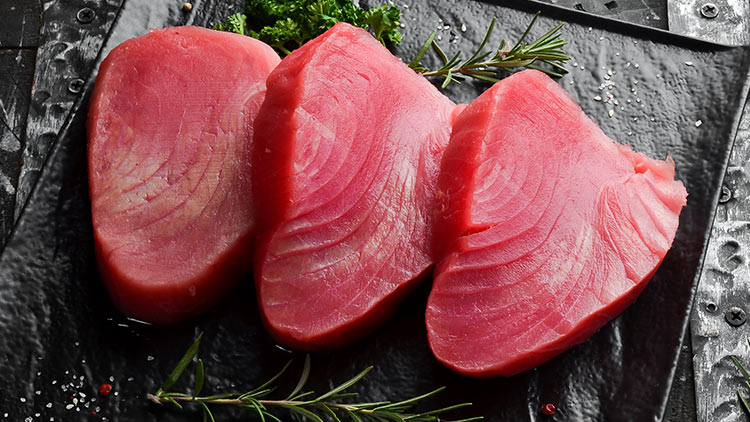 Drei rohe Thunfischstücke auf schwarzer Platte mit Rosmarinzweigen, Petersilie und einer angeschnittenen roten Zwiebel.