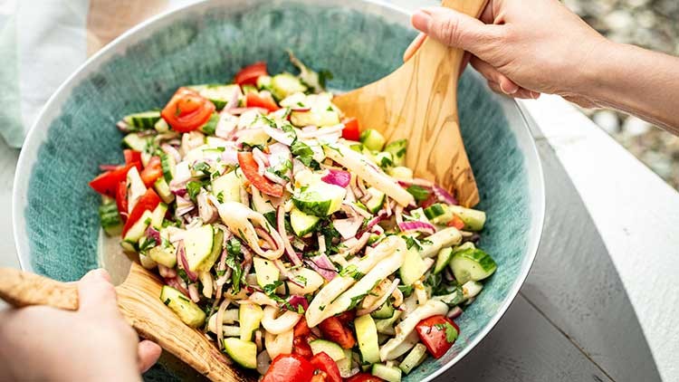Eine große Salatschüssel mit Gurken, Tomaten, Zwiebeln und Kräutern wird mit zwei Holzsalatlöffeln gemischt.