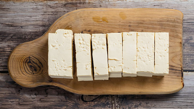 Acht rechteckige Stücke weißer Käse liegen auf einem hellbraunen Holzbrett auf einer rustikalen Holzoberfläche.
