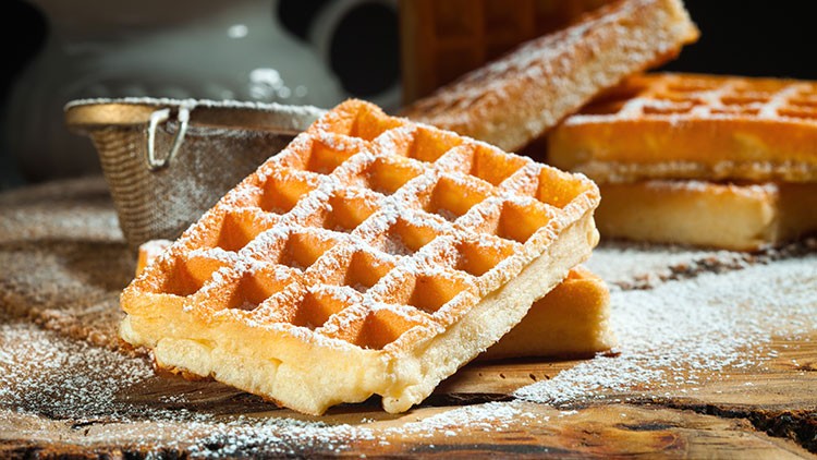 Frische, goldbraune Waffeln mit Puderzucker auf einem rustikalen Holzuntergrund, im Hintergrund ein Sieb.