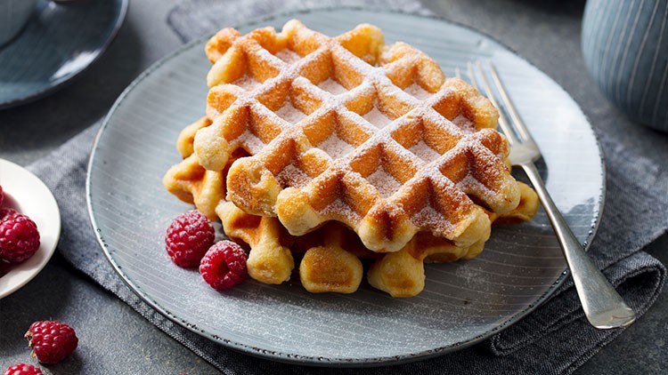 Zwei goldbraune Waffeln mit Puderzucker liegen auf einem grauen Teller, daneben frische Himbeeren und eine Gabel.