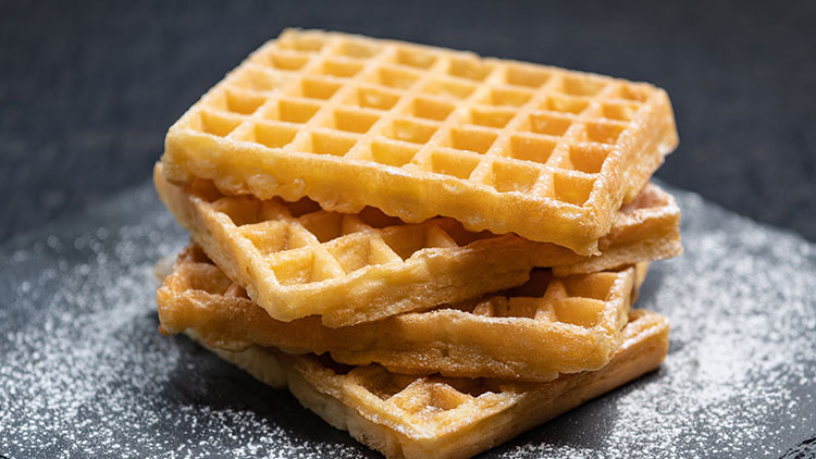 Ein Stapel goldbrauner Waffeln liegt auf einer dunklen Schieferplatte und ist mit Puderzucker bestreut.