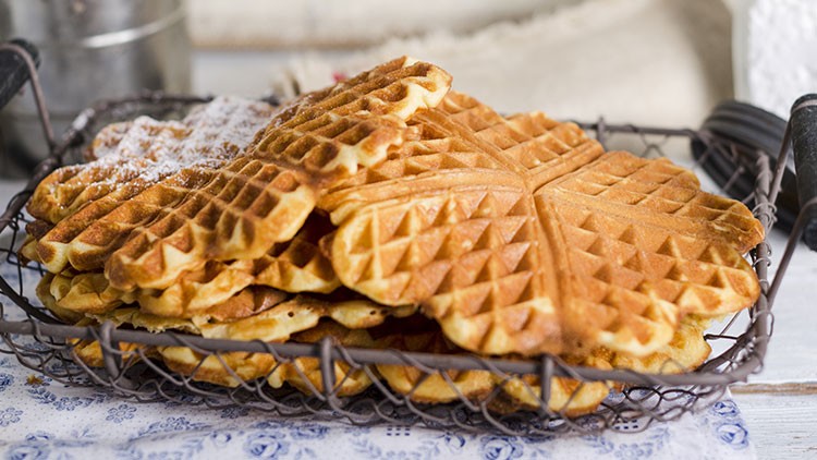 Mehrere goldbraune, herzförmige Waffeln liegen gestapelt in einem Drahtkorb, eine Waffel ist mit Puderzucker bestreut.