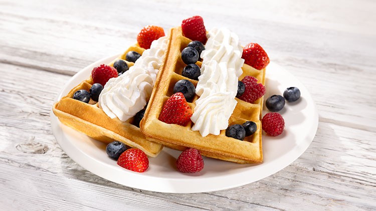 Zwei Waffeln mit Sahne, Himbeeren und Blaubeeren auf weißem Teller, Hintergrund aus hellem Holz.