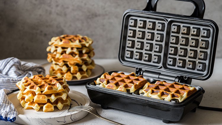 Geöffneter Waffelautomat mit zwei gebackenen Waffeln, daneben Teller mit gestapelten Waffeln und blau-weißem Tuch.