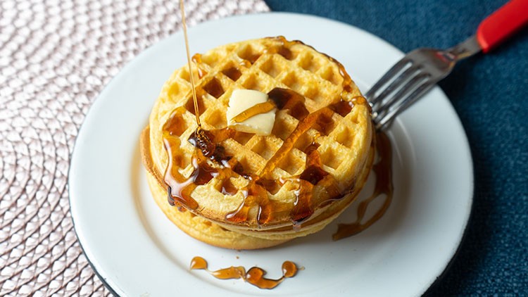 Zwei gestapelte Waffeln mit einem Butterstück, übergossen mit Sirup, auf einem weißen Teller mit einer Gabel am Rand.