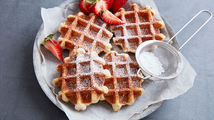 Vier belgische Waffeln mit Puderzucker, frischen Erdbeeren und einem Sieb auf einem Teller mit Backpapier.