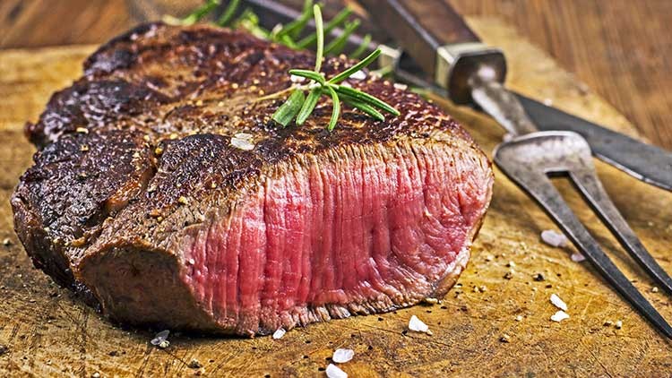 Ein aufgeschnittenes, medium gebratenes Steak liegt auf einem Holzbrett, dekoriert mit Rosmarinzweigen und grobem Salz.