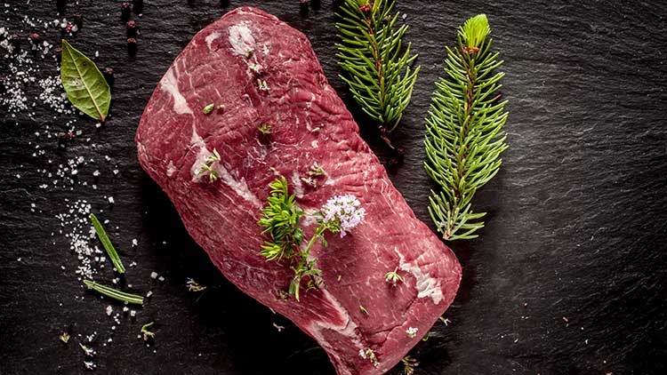 Ein Stück rohes Rindfleisch mit Kräutern dekoriert, daneben liegen Tannenzweige, Lorbeerblätter und feine Gewürze auf schwarzem Untergrund.