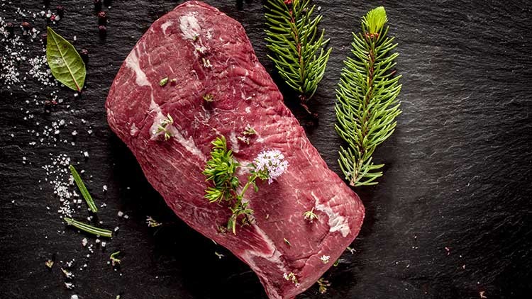 Ein rohes Stück Fleisch liegt auf einer dunklen Schieferplatte, dekoriert mit Kräutern, Blüten, Salz und Tannenzweigen.
