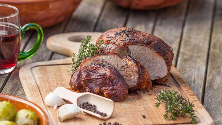 Ein gebratener Fleischbraten, angeschnitten und dekoriert mit frischem Thymian, liegt auf einem Holzbrett mit Knoblauch und Pfeffer.