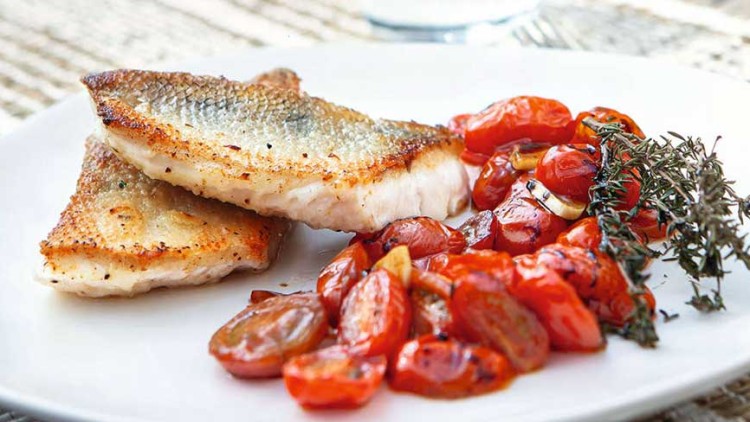 Zwei gebratene Fischfilets mit gebräunter Haut, geschmorte Kirschtomaten und frische Kräuter auf einem weißen Teller.