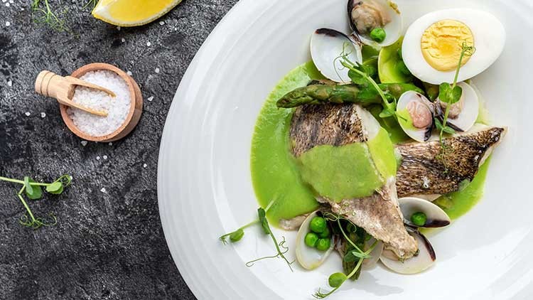 Gegrilltes Fischfilet mit grüner Soße, Spargel, Erbsen, Muscheln und einem halbierten Ei auf einem weißen Teller.