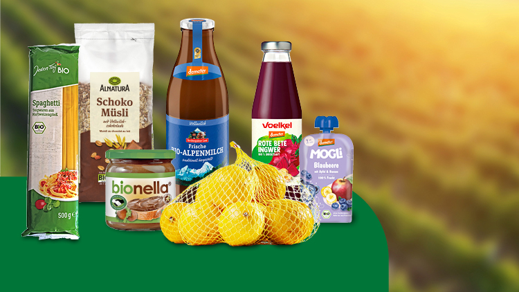 Verschiedene Bio-Lebensmittel wie Spaghetti, Müsli, Alpenmilch, Rote-Bete-Saft, Zitronen und Blaubeer-Fruchtmus vor einem Feld.