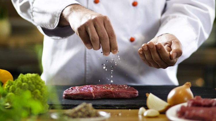 Eine Person in Kochjacke würzt ein rohes Stück Fleisch mit grobem Salz, daneben liegen Gemüse wie Zwiebeln und Brokkoli.