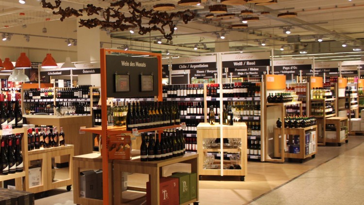 Weinabteilung mit Regalen voll Flaschen, orangem Schild „Wein des Monats“ und Beschriftungen wie „Chile/Argentinien“ und „Weiß/Rosé“.
