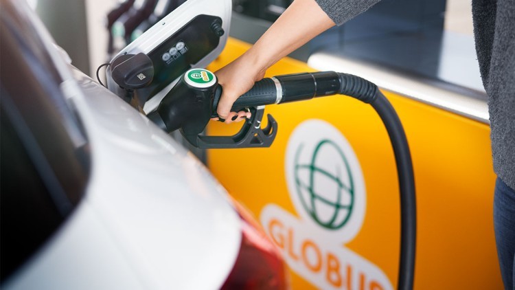 Eine Hand hält eine Zapfpistole an einem weißen Auto, im Hintergrund das orange GLOBUS-Logo.